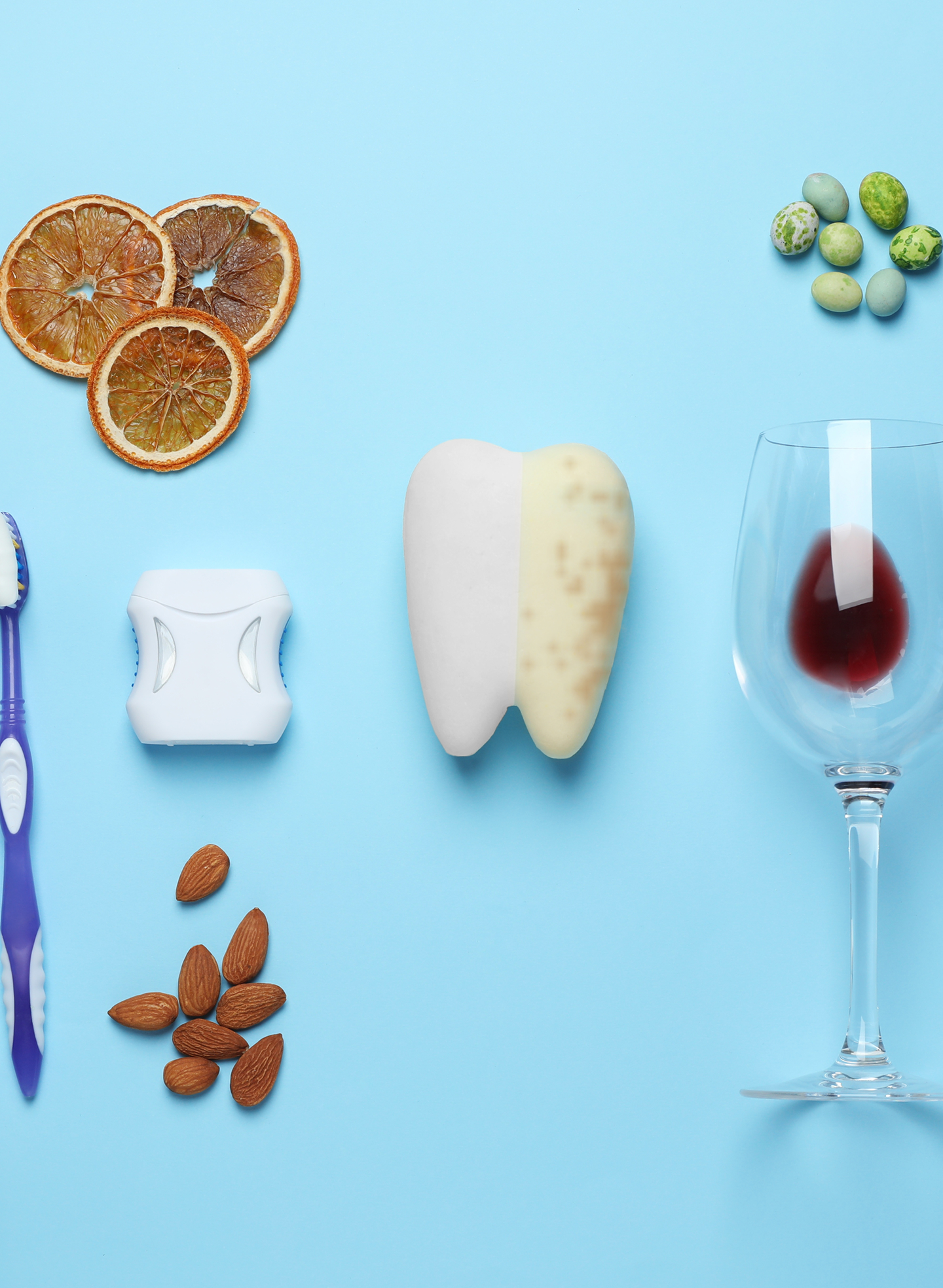 A tooth model displays one side white and the other stained, illustrating different types of teeth stains. Surrounding it are stain-causing items like red wine, dried citrus, and nuts, along with a toothbrush and floss for prevention. This image highlights common causes of tooth discoloration and the importance of oral care, aligned with Aspen Dental’s focus on healthy smiles.