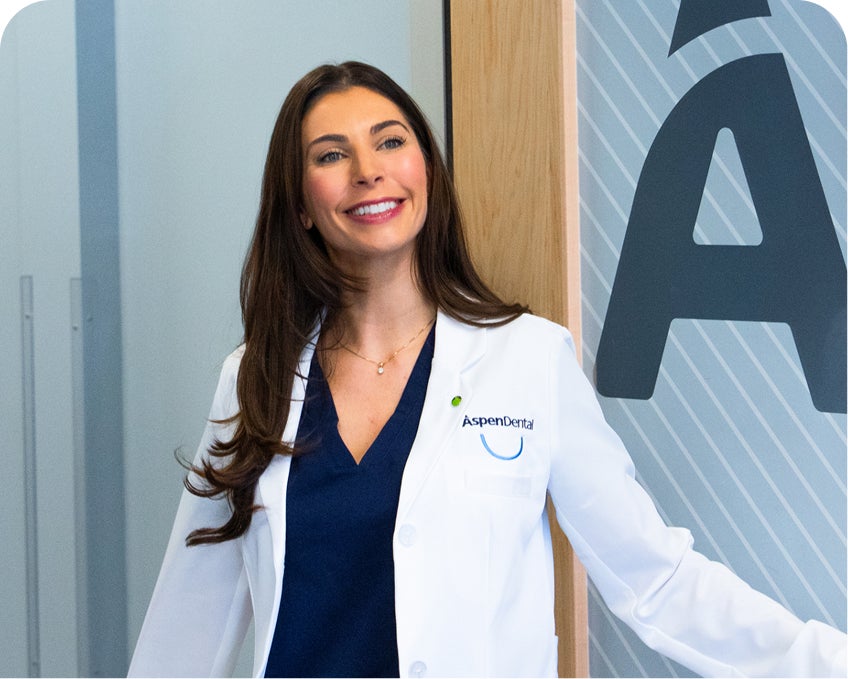Smiling Aspen Dental professional welcomes patients at the office door, representing the first step in enrolling in the Aspen Dental Savings Plan for affordable, no-judgment dental care.