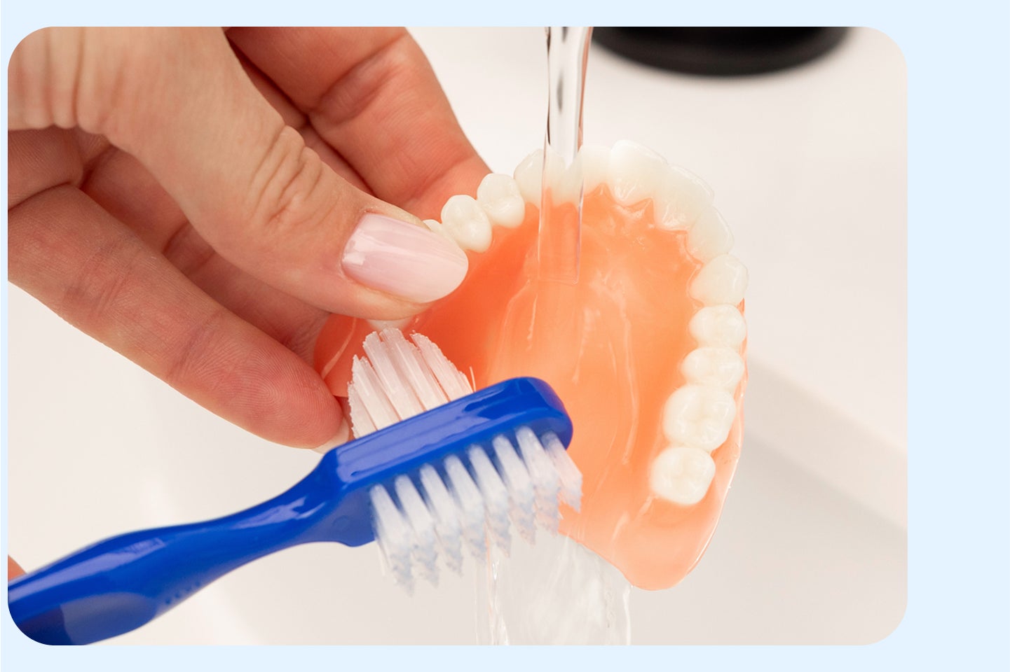 Brushing an upper denture under running water with a soft toothbrush as part of daily denture care routine.