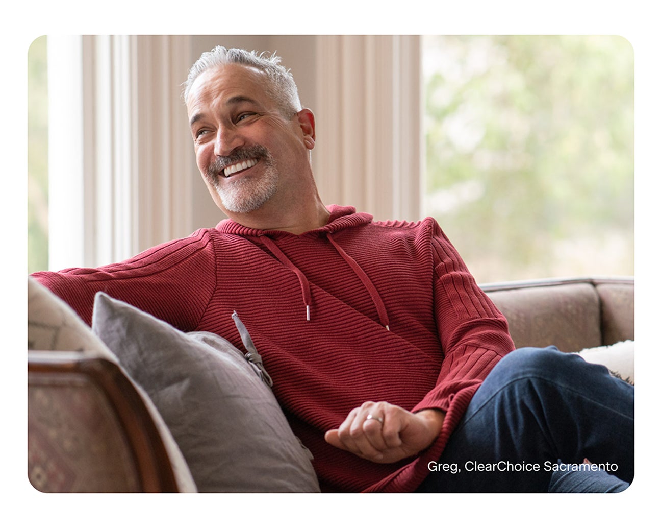Greg, a ClearChoice Sacramento patient, smiling confidently while relaxing on a sofa in a red sweater, showcasing the results of dental implants and improved quality of life.