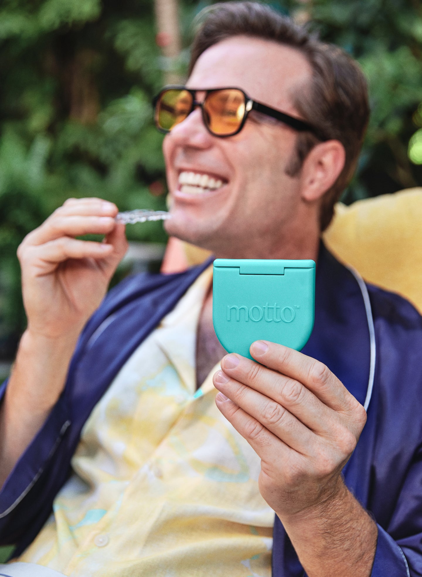 A man wearing sunglasses and a blue robe is holding Motto clear aligners