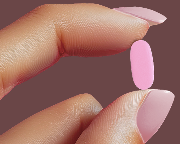 A hand with pink painted nails holds a bright pink pill against a dark background