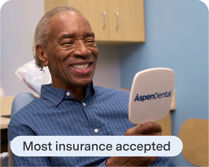 An Aspen Dental patient seated in a treatment room smiles while looking into an Aspen Dental mirror