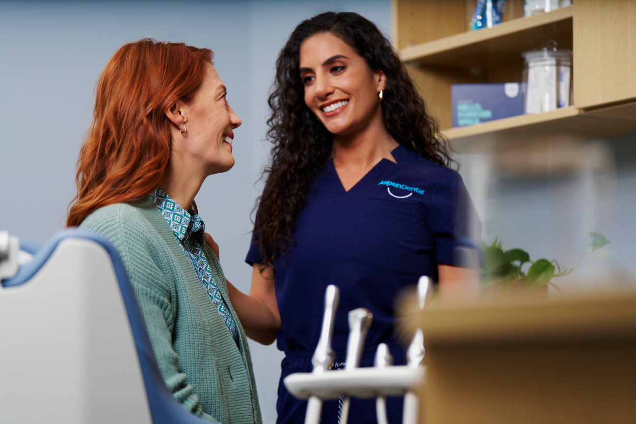  A smiling Aspen Dental staff member in uniform providing reassurance to a patient seated in a treatment room, creating a welcoming and professional atmosphere.