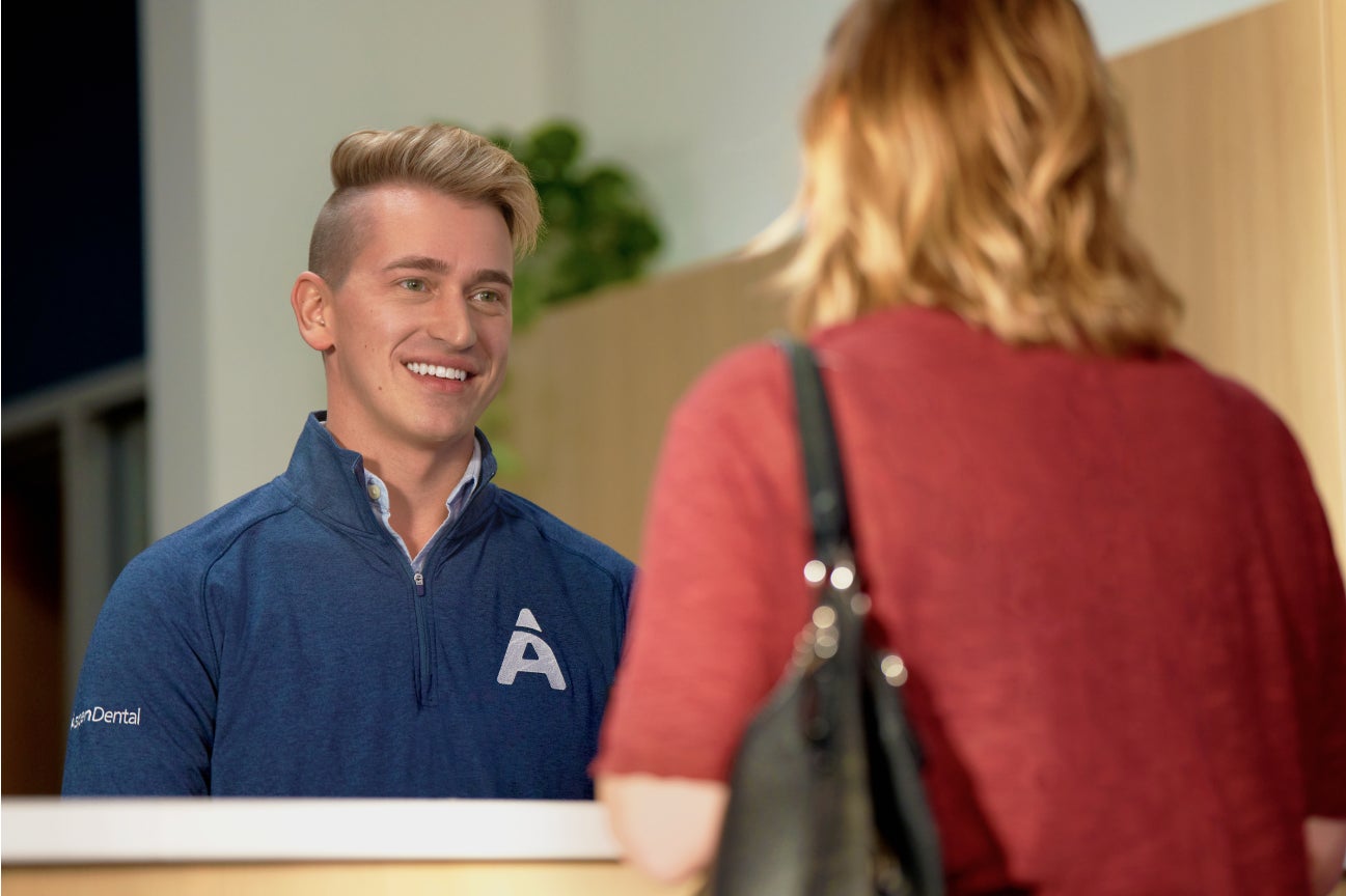 A friendly receptionist at Aspen Dental assisting a patient at the front desk, emphasizing welcoming service and consultation opportunities for tooth cap treatments.