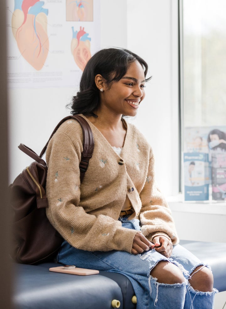 A woman with a backpack is sitting in a doctor's office and smiling.