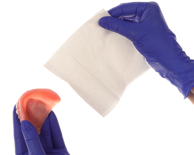  A person wearing gloves drying dentures with a paper towel to remove food particles and saliva. Aspen Dental recommends proper preparation for denture hygiene.