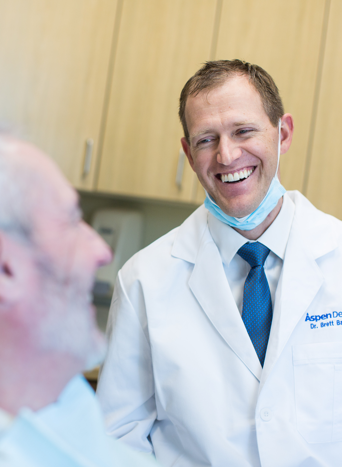 An Aspen Dental dentist in a white coat and blue tie smiles while discussing oral surgery costs with a patient. The consultation takes place in a modern dental office, emphasizing affordable and personalized care options.