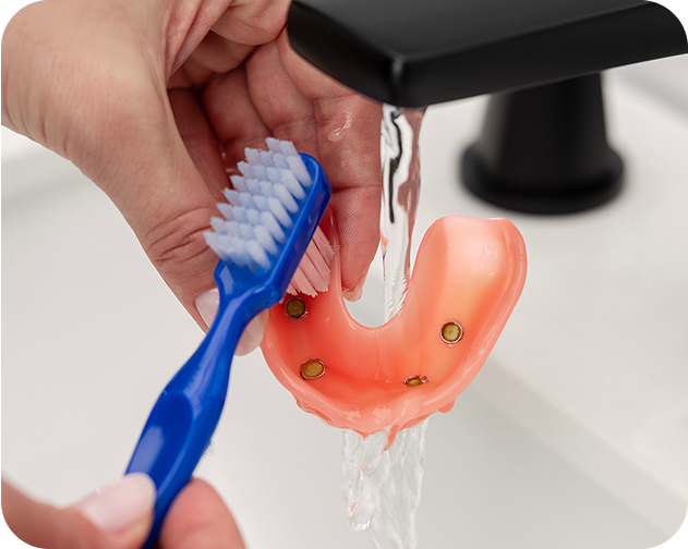 Cleaning an implant denture under running water using a blue toothbrush – Aspen Dental oral hygiene care.