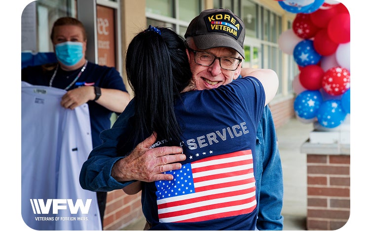 An older man wearing a "Korea Veteran" cap receives a warm hug from an Aspen Dental team member outside a dental office, with patriotic balloon decorations in the background. The moment reflects gratitude, support, and appreciation for veterans.