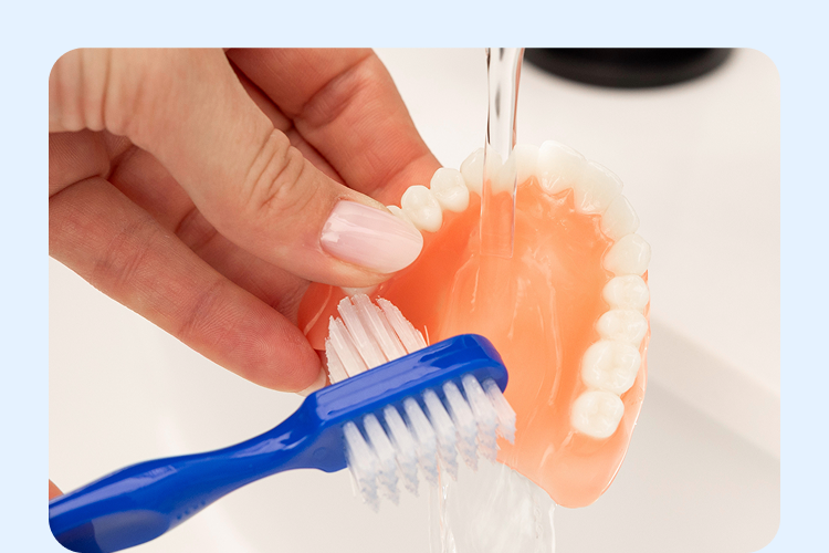 Brushing an upper denture under running water with a soft toothbrush as part of daily denture care routine.