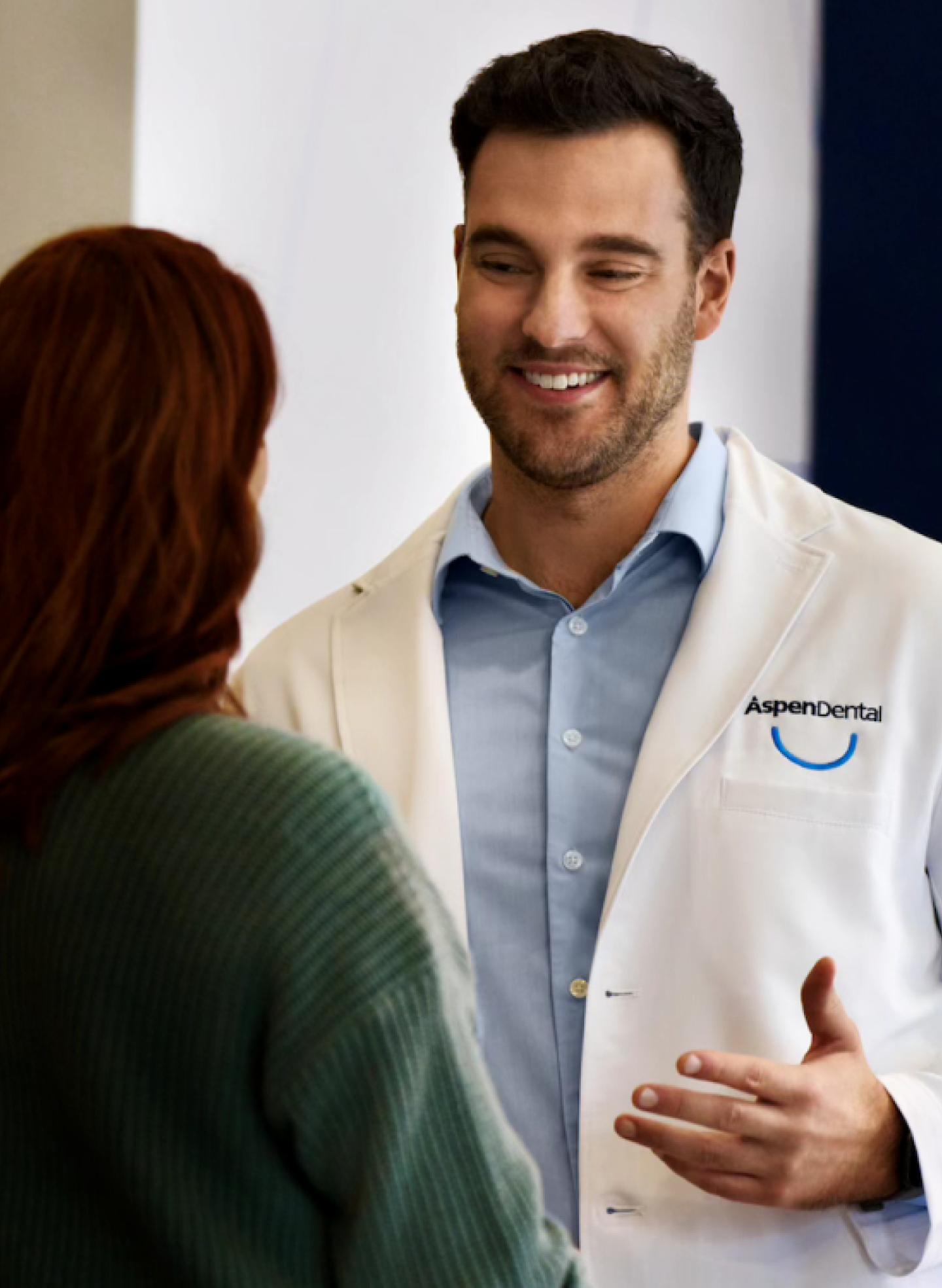 A friendly Aspen Dental dentist in a white coat smiling and consulting with a patient in a green sweater during an appointment.