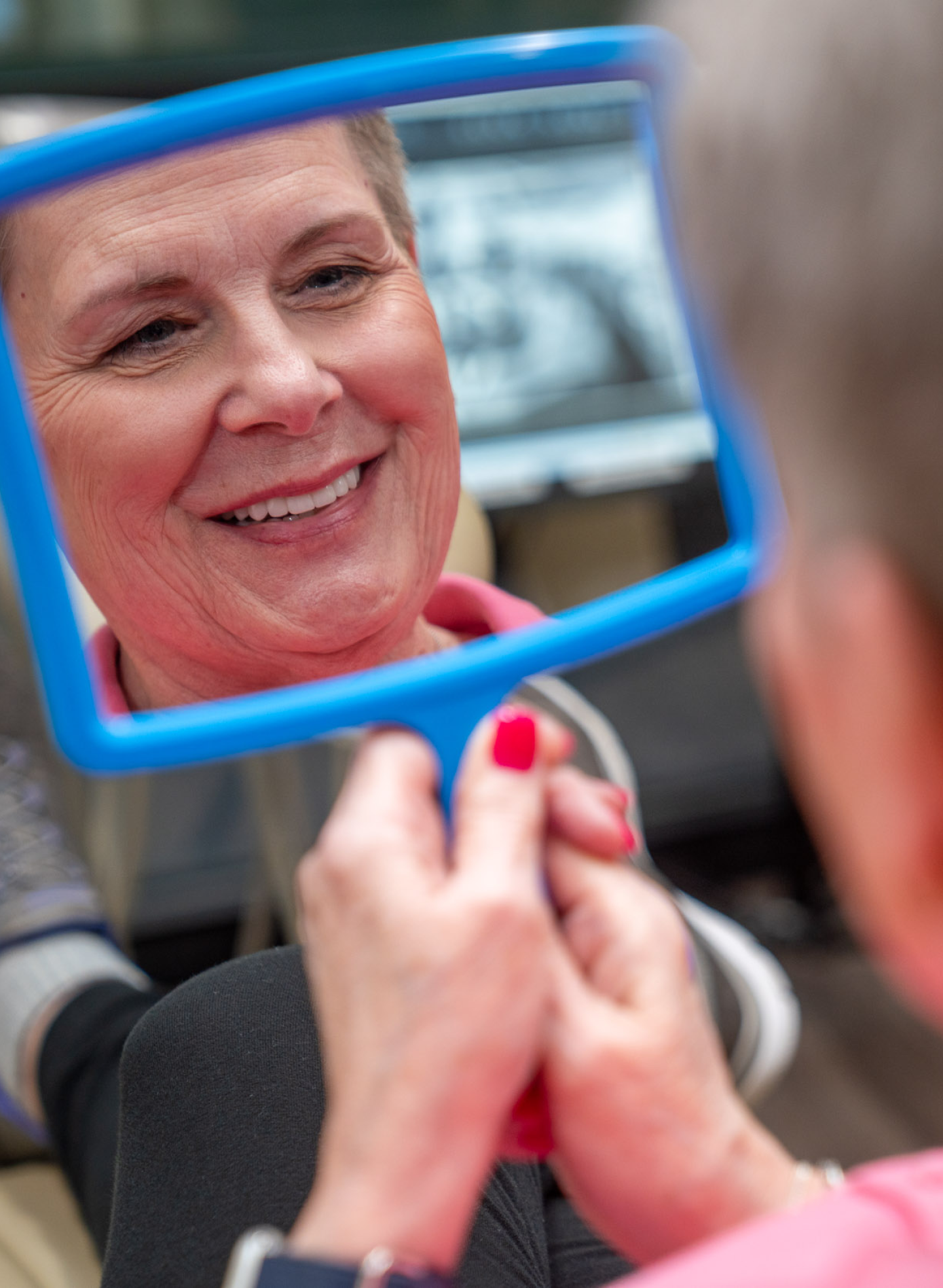 A smiling older woman looking at her reflection in a handheld mirror, showcasing her dental results, representing the outcomes of a dental implant procedure at ClearChoice.