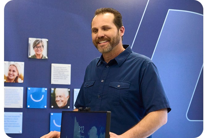 A joyful man with a beard and a blue shirt stands at the front desk of an Aspen Dental office to inquire about denture treatment. The image conveys excitement, symbolizing the patient's enthusiasm for learning about the process it takes to get dentures.