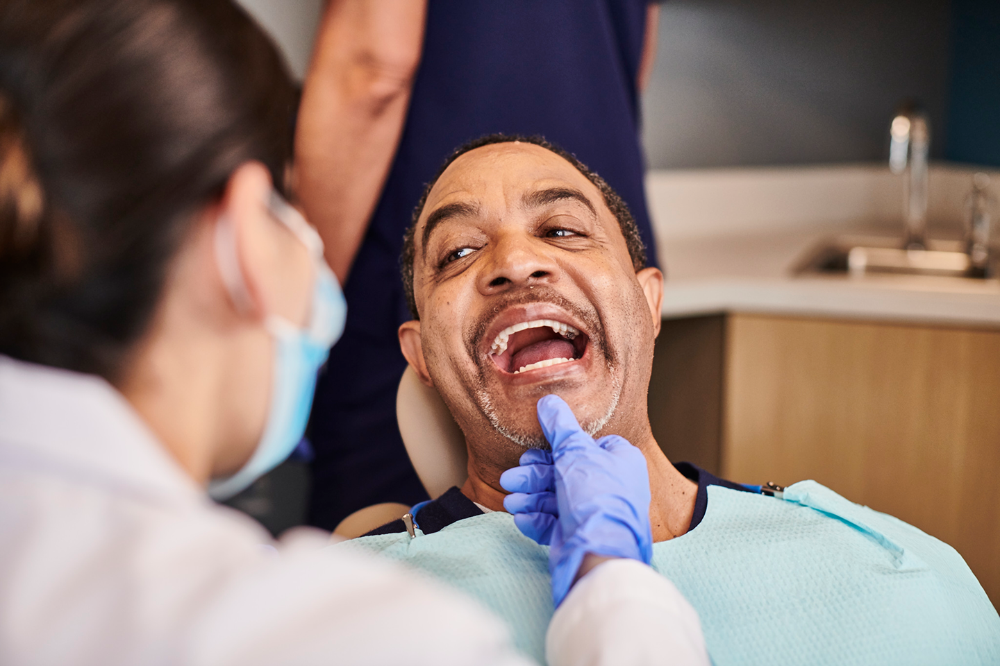 A male patient sits in a dental chair, smiling while a dentist wearing a mask and gloves examines his gums. The professional checkup focuses on gum health and detecting signs of gum disease. Aspen Dental provides expert care to diagnose and treat oral health issues.