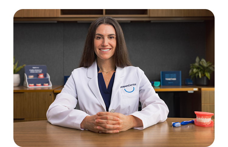 Aspen Dental provider seated at desk with denture model and toothbrush for patient care education.