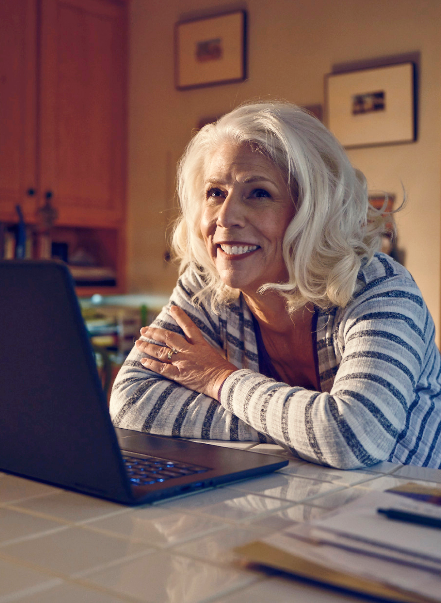 Smiling senior woman with white hair sitting at a kitchen table using a laptop, researching the pros and cons of dental implants.