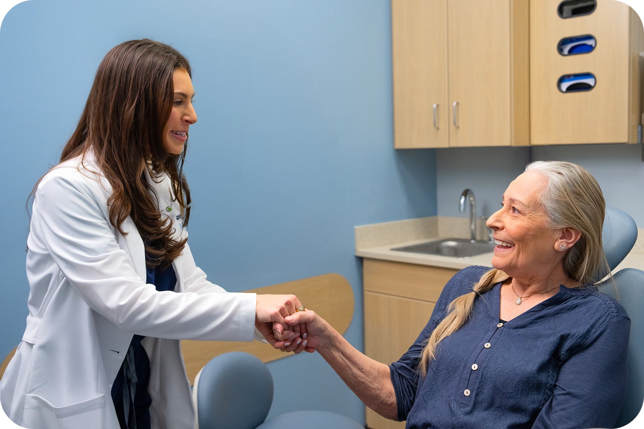 An Aspen Dental provider warmly greets an older woman in a treatment chair during her first visit for dentures, both smiling in a clean exam room.