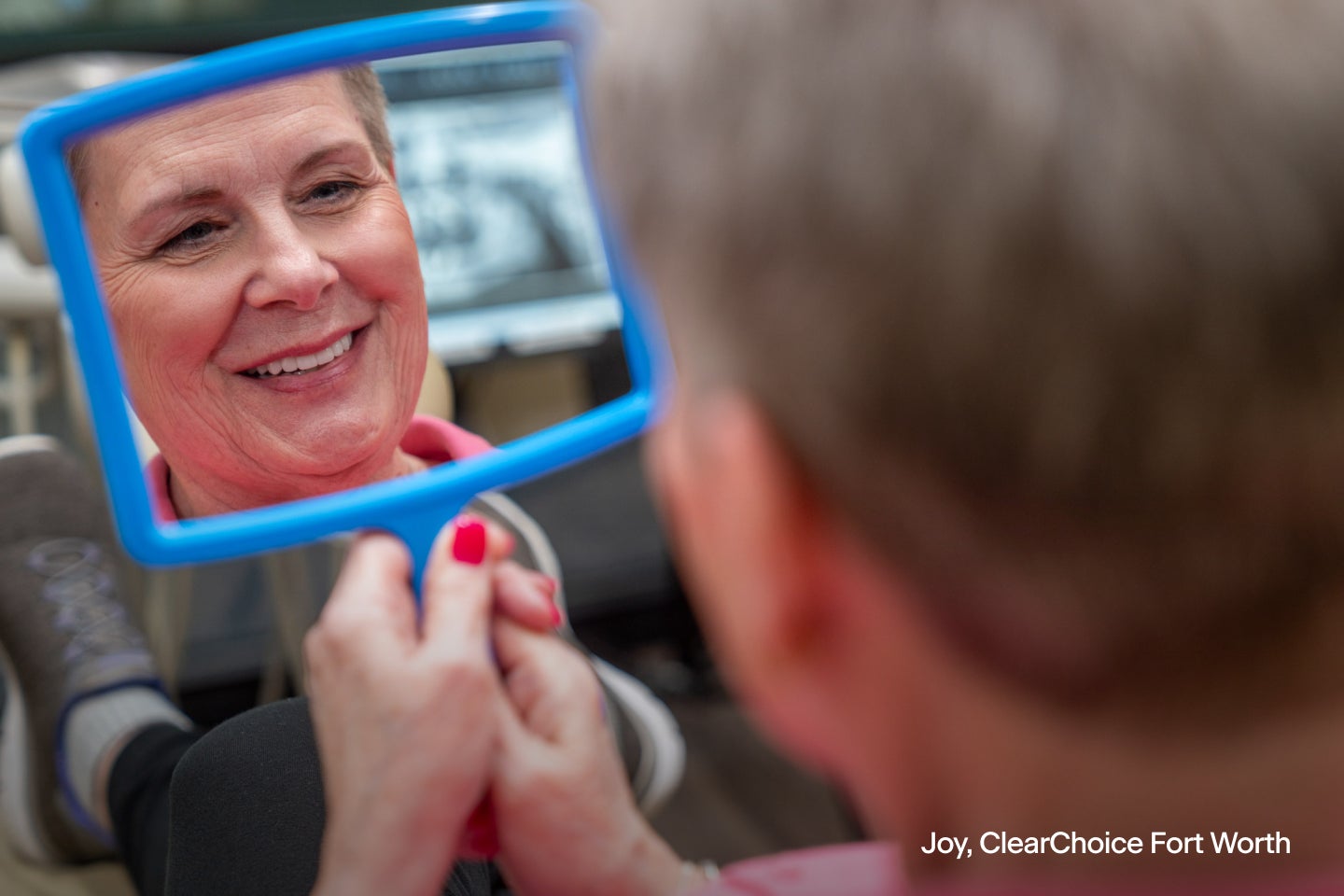 An image of Joy, a ClearChoice dental implant patient from Fort Worth, smiling into a blue handheld mirror.