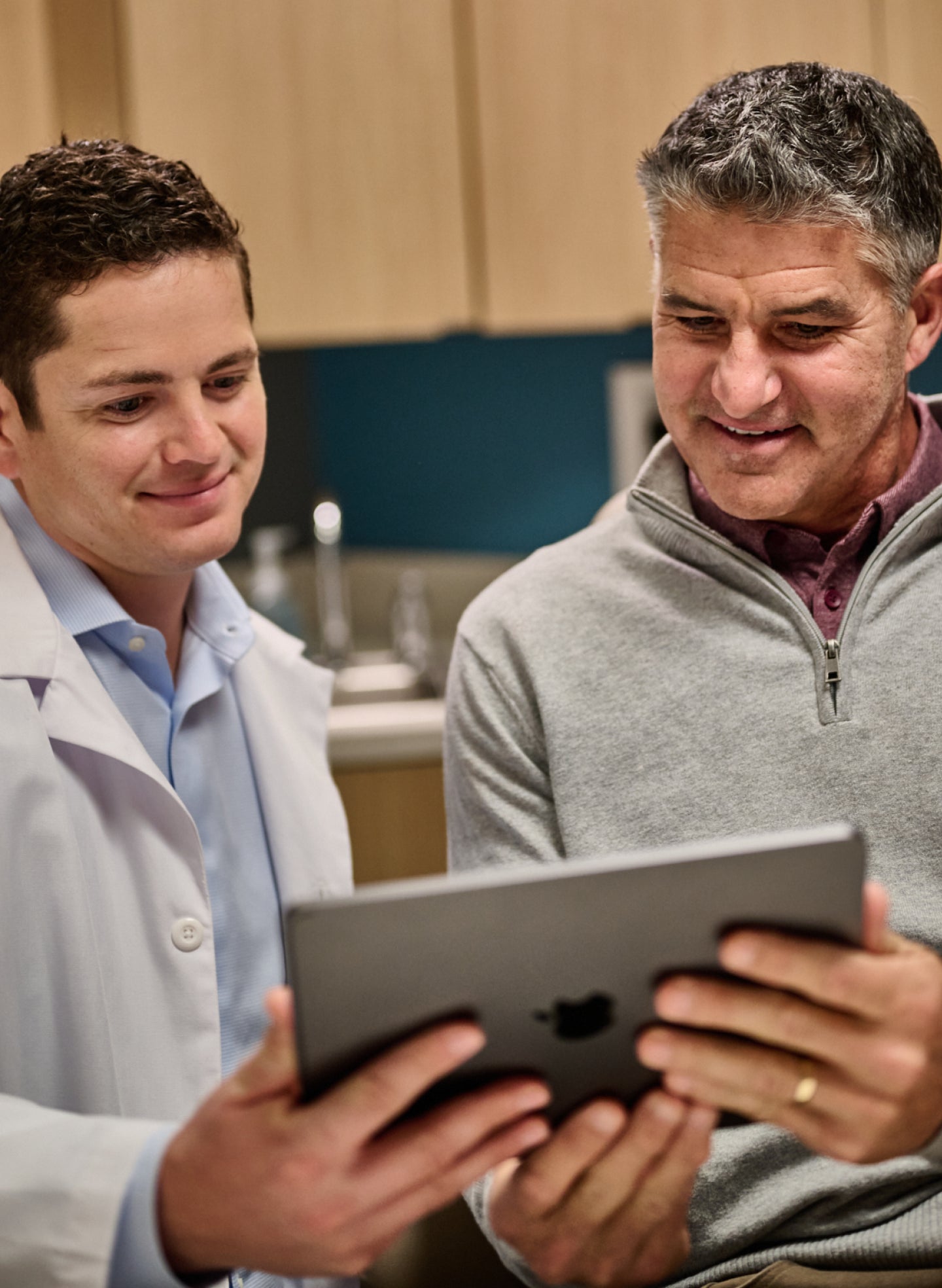 A photo of an Aspen Dental dentist with a white coat and light blue button up consulting with his patient, who is wearing a light gray quarter zip, while both are holding an iPad and smiling.