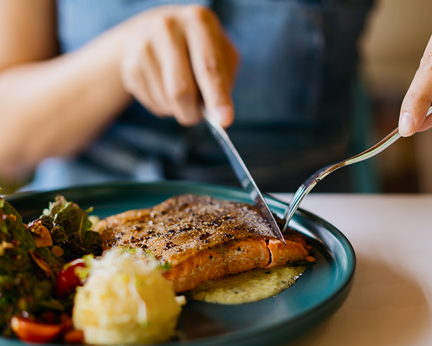  A close-up of a person cutting into a nutritious meal with grilled fish, vegetables, and mashed potatoes, highlighting the importance of a balanced diet for strong teeth and gums. Aspen Dental encourages healthy eating for better oral health.