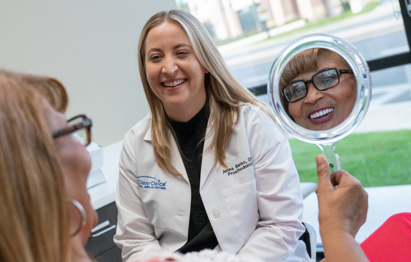 An older woman smiles into a handheld mirror while sitting with a ClearChoice dental specialist, reflecting the positive AARP dental care experience and showing her new dental implants.