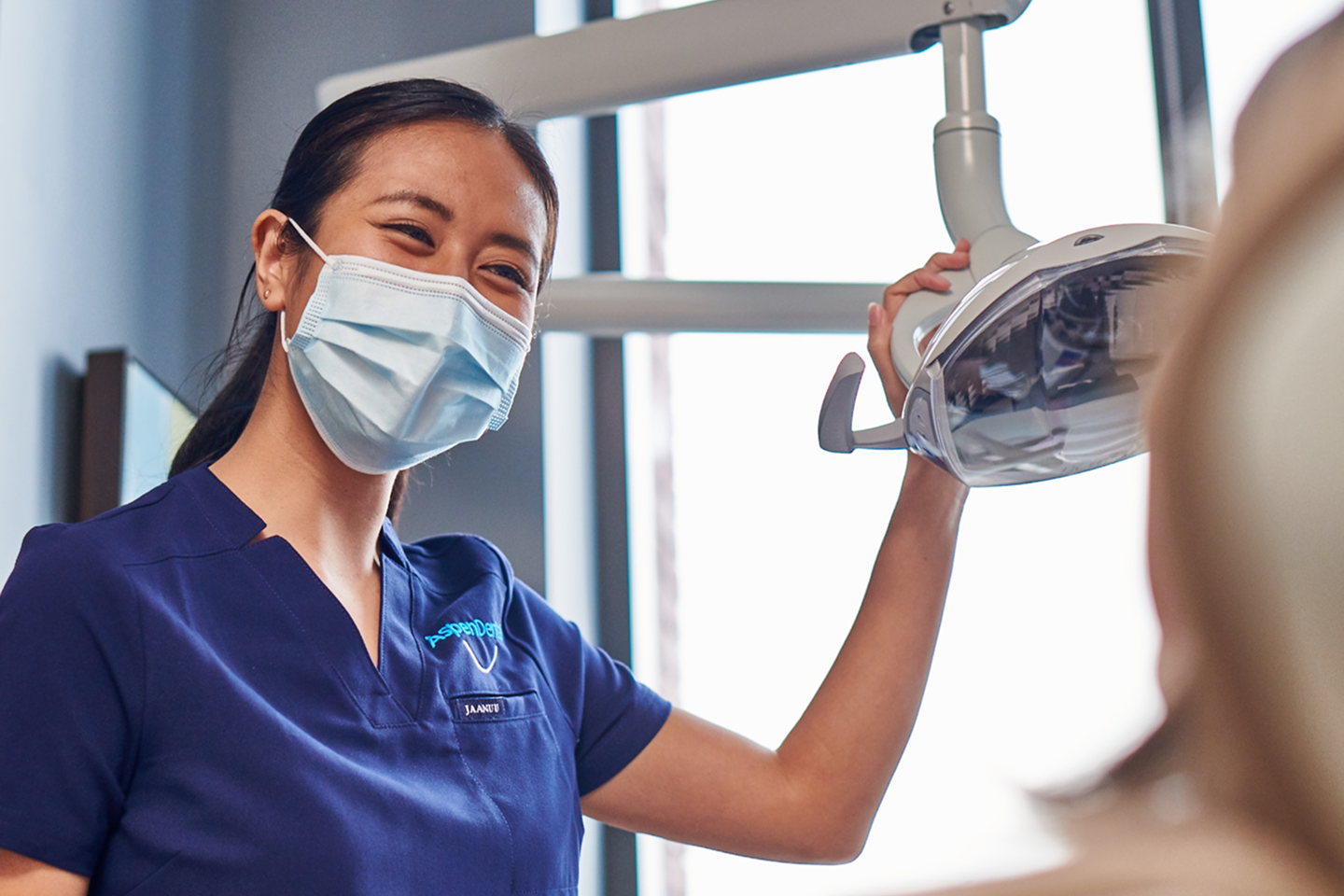 A friendly Aspen Dental professional wearing a mask adjusts an overhead light while preparing for a patient's dental exam. Aspen Dental prioritizes compassionate and expert dental care for every patient.