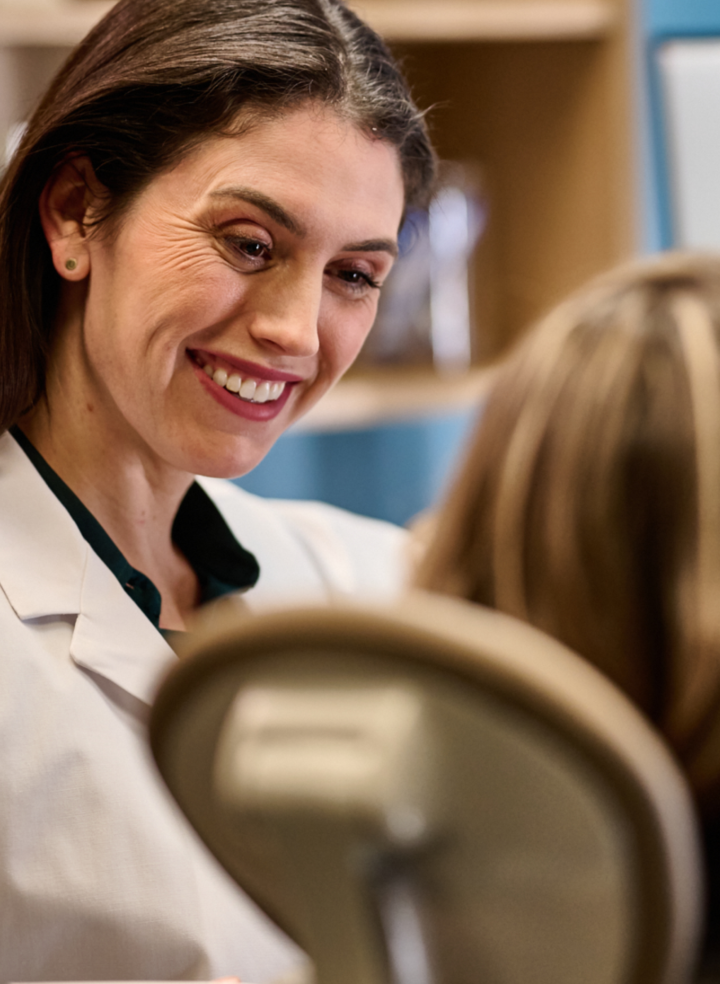 A female dentist in a white coat smiling and talking to a patient, who is seated with their back to the camera. Medical equipment is visible in the background.