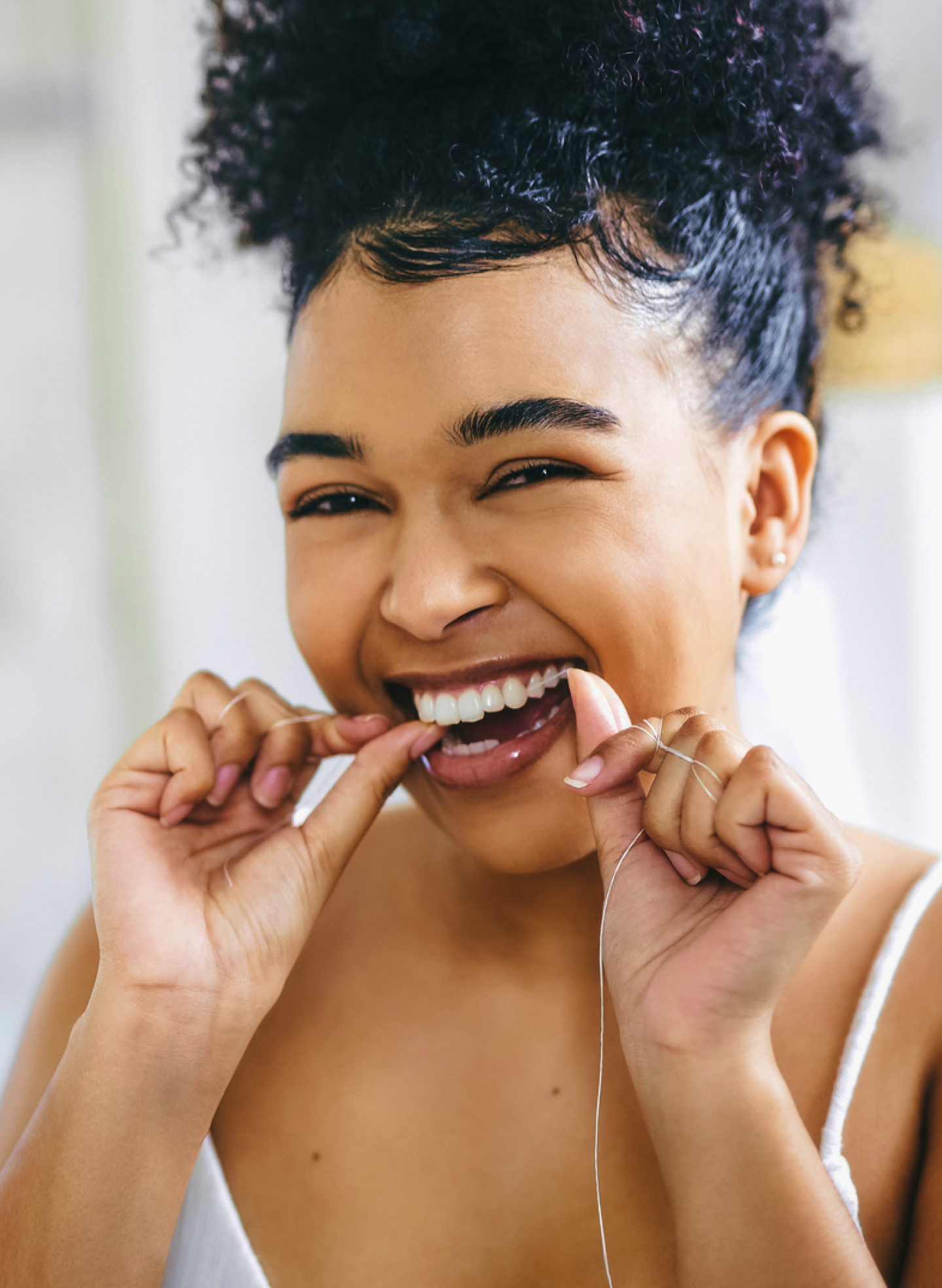 A woman with curly hair smiles brightly while flossing her teeth, promoting good oral hygiene. She is wearing a white tank top, emphasizing a clean and fresh dental care routine associated with Aspen Dental.