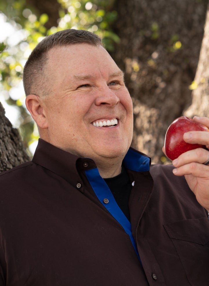 A smiling man holding a red apple outdoors, showcasing bright teeth after receiving molar dental implants from ClearChoice, with natural greenery in the background.