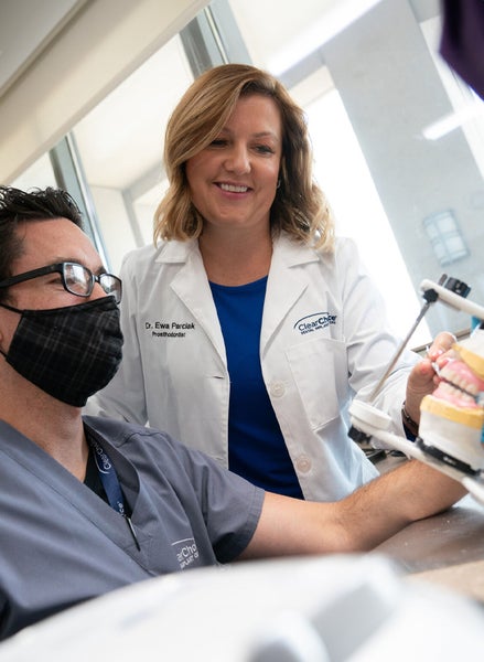 A man wearing a mask is sitting at a table working on a set of dental implants with a woman in a lab coat helping