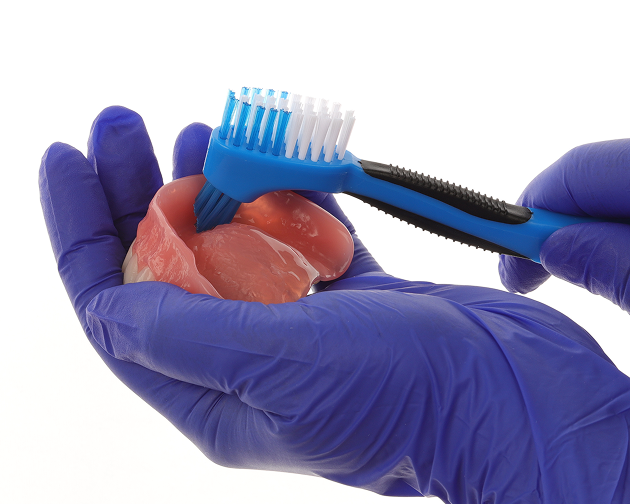A person in gloves brushing dentures under lukewarm water with a soft-bristled toothbrush. Aspen Dental promotes gentle cleaning for denture maintenance.