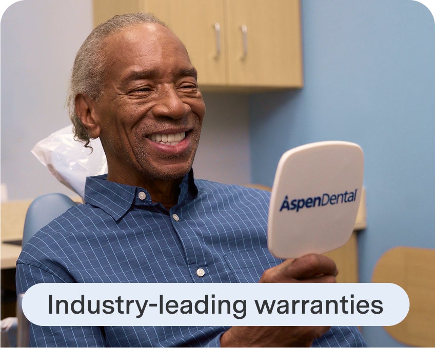 Aspen Dental patient smiles while looking at his new dentures in the mirror, highlighting leading-industry warranties.