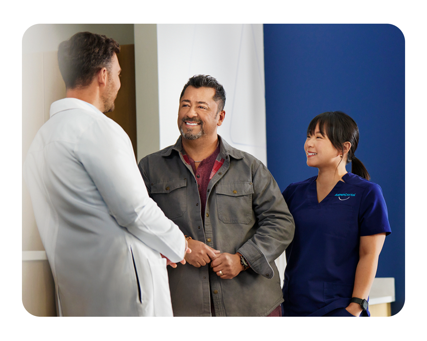 A smiling Aspen Dental patient speaks with a dentist and a team member in a welcoming office. The image highlights a supportive consultation process for dental implants and tooth replacement options.