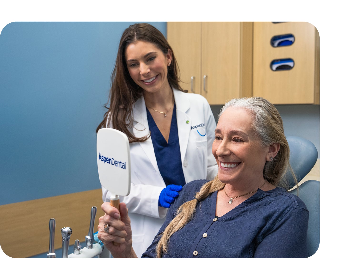 An older woman smiles while looking into a handheld mirror after receiving dentures, as an Aspen Dental professional stands beside her in a treatment room.