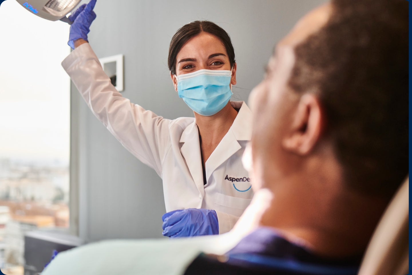  An Aspen Dental dentist wearing a mask and gloves adjusts the overhead light while preparing a patient for a veneer procedure in a modern dental office setting.