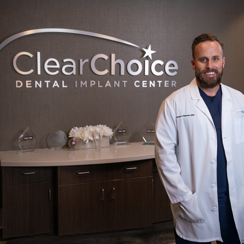 A ClearChoice dental professional in a white coat standing beside the ClearChoice Dental Implant Center reception desk, welcoming patients to a warm and professional environment.