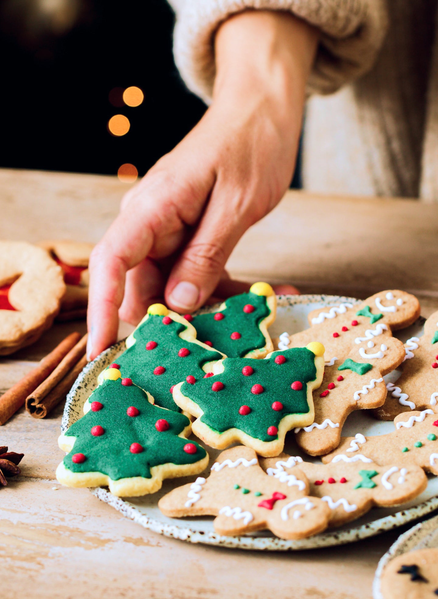  A plate of festive holiday cookies featuring green Christmas tree-shaped cookies with red icing dots and gingerbread cookies decorated with white icing, with a hand reaching for one of the tree cookies.