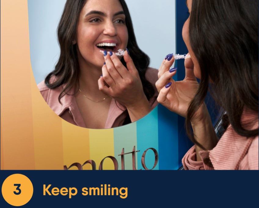 Smiling patient in a Motto dental setting holding a clear aligner near her mouth as she looks into a mirror, preparing to place the aligner while dressed casually in a warm-toned shirt.