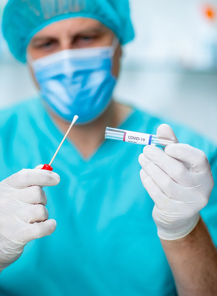 A doctor wearing a mask and gloves is holding a swab and a test tube that says COVID-19 on it