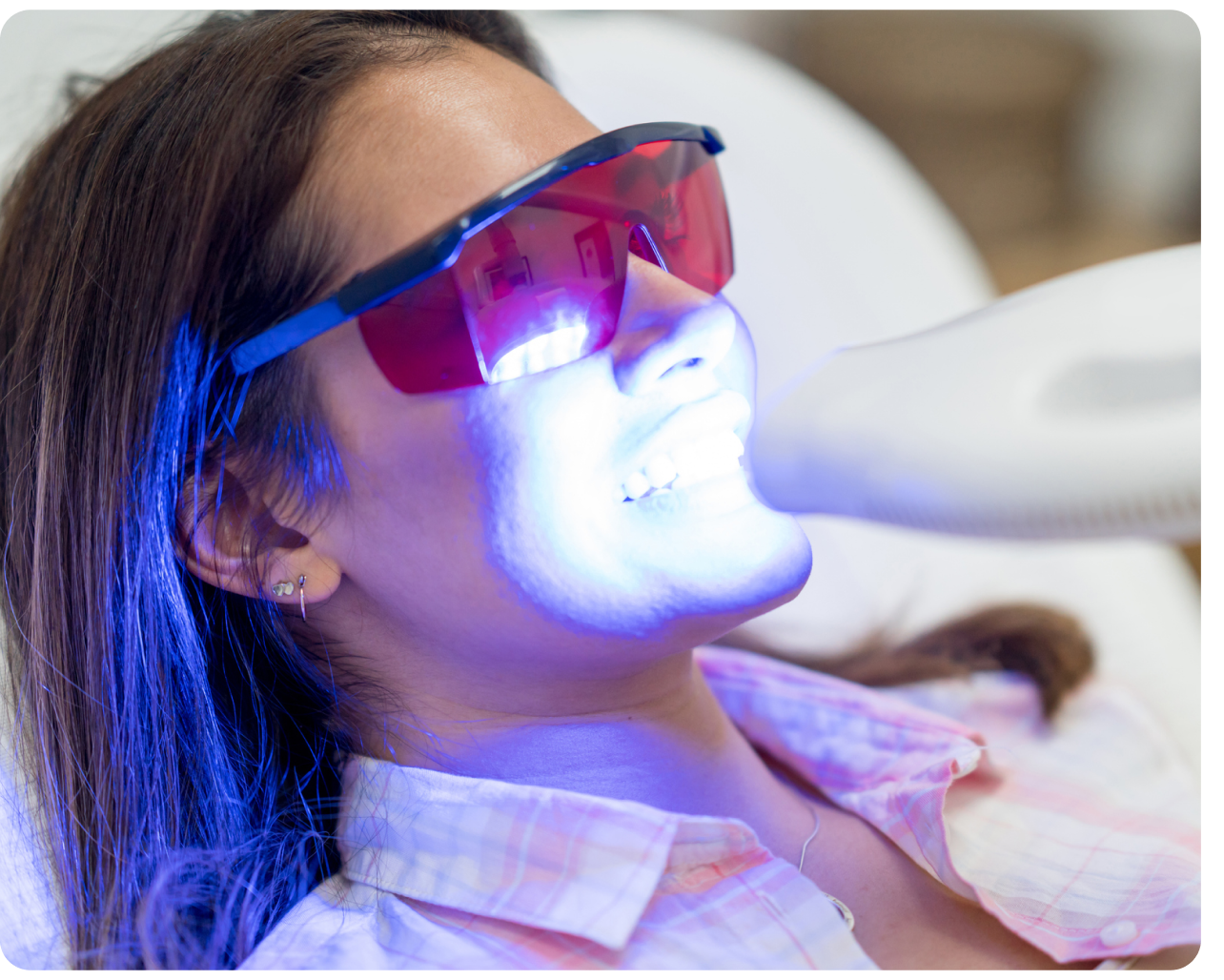 Patient undergoing an in-office teeth whitening procedure at Aspen Dental, wearing protective glasses as a blue LED light activates the whitening agent on their teeth.