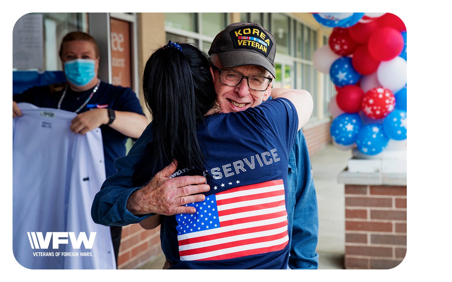 An older man wearing a "Korea Veteran" cap receives a warm hug from an Aspen Dental team member outside a dental office, with patriotic balloon decorations in the background. The moment reflects gratitude, support, and appreciation for veterans.