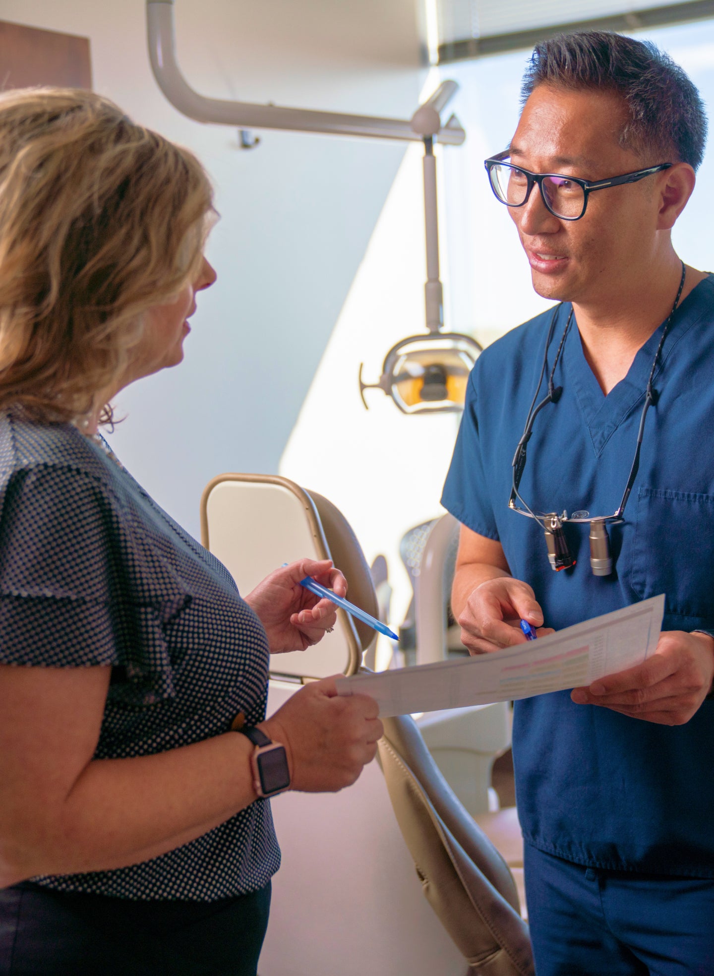 A ClearChoice dental professional in blue scrubs discussing dental implant options with a patient in a modern dental office. The dentist is holding a treatment plan and explaining details to the patient.
