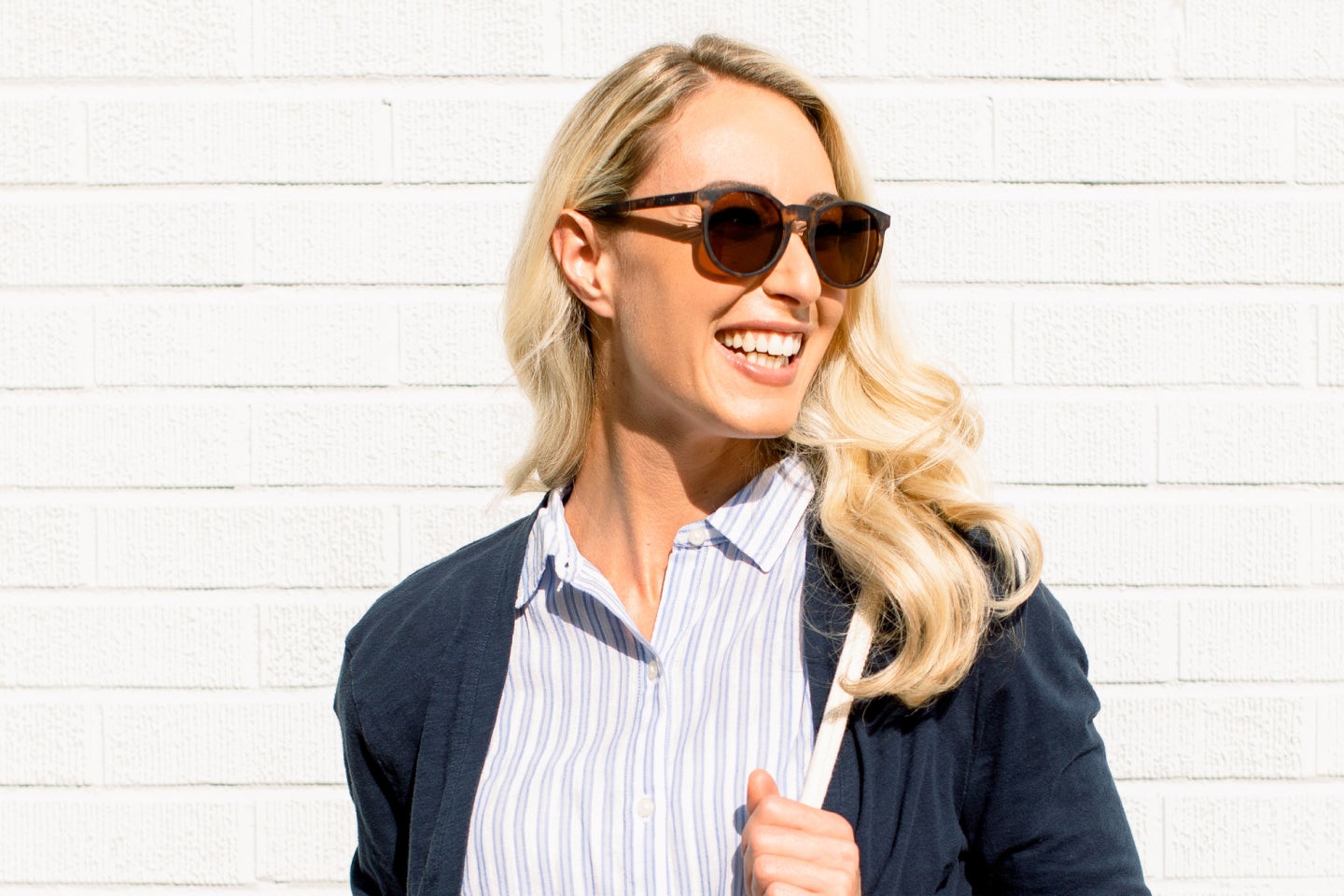  A confident woman smiling brightly while wearing sunglasses, standing against a white brick wall in natural sunlight, embodying the confidence and joy inspired by Motto clear aligners and their transformative results.