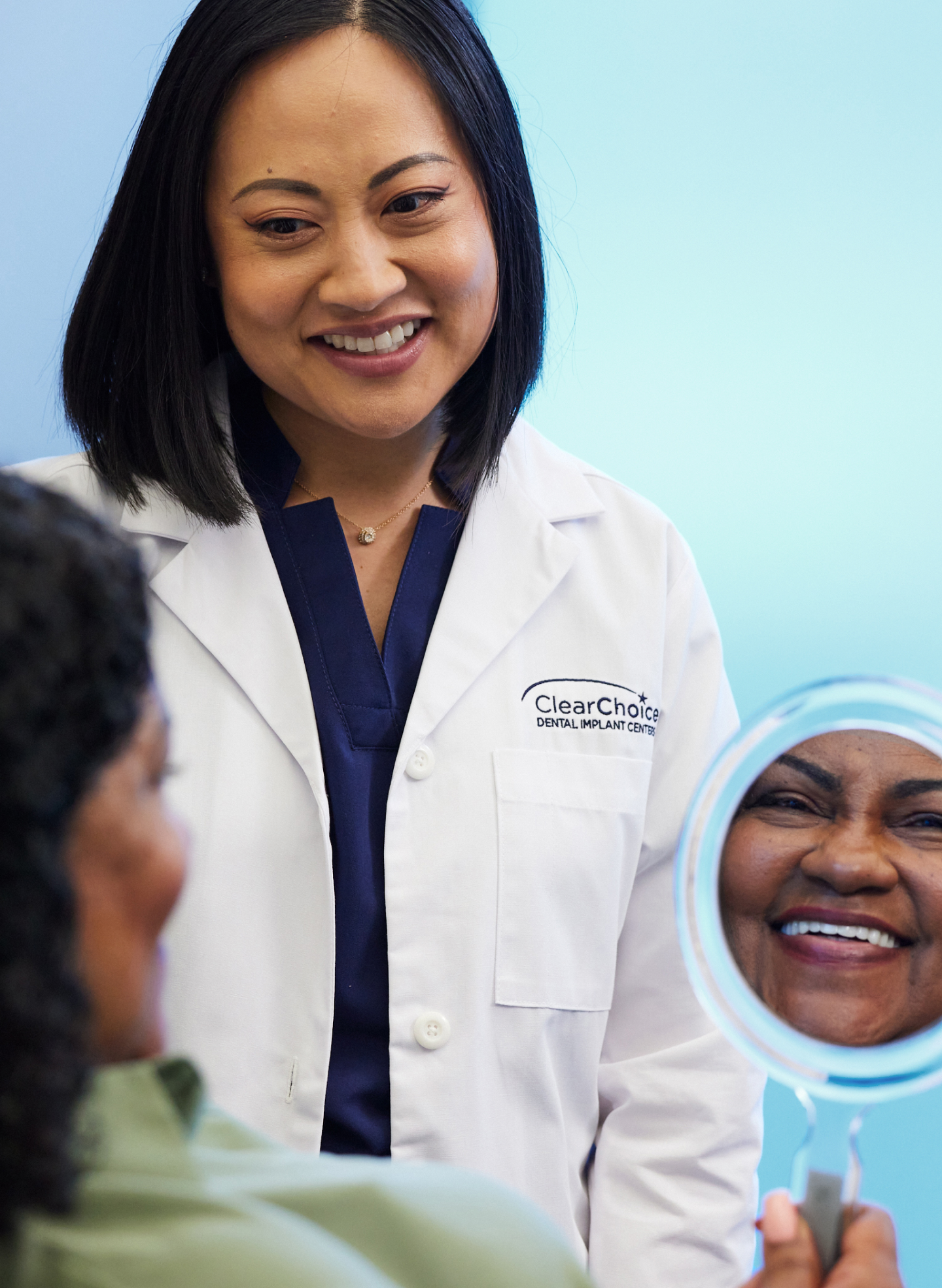 ClearChoice dentist smiling with a patient who is viewing her smile in a handheld mirror, reflecting satisfaction with dental care in a welcoming clinic environment.