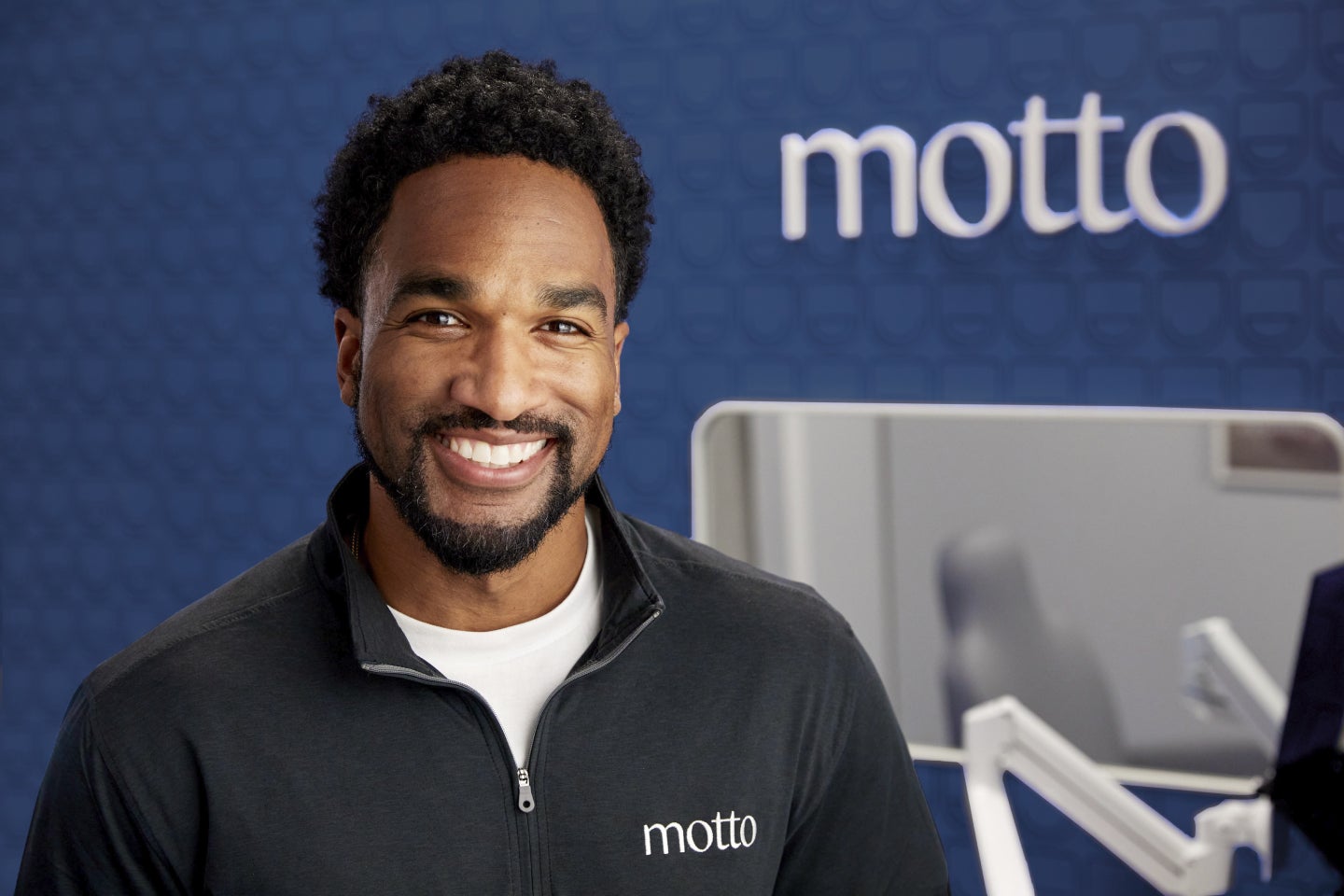 A smiling Motto dental professional wearing a Motto-branded jacket, standing confidently in a modern dental office with the Motto logo prominently displayed in the background, representing the expertise and care provided by Motto clear aligners.