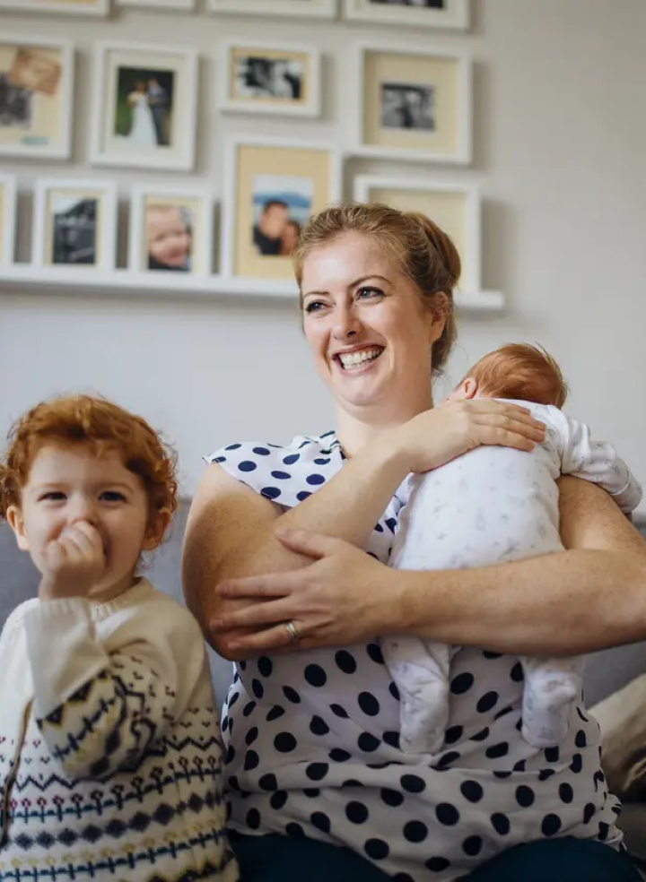 A woman is sitting on a couch holding a baby while her other child stands in front of her. 