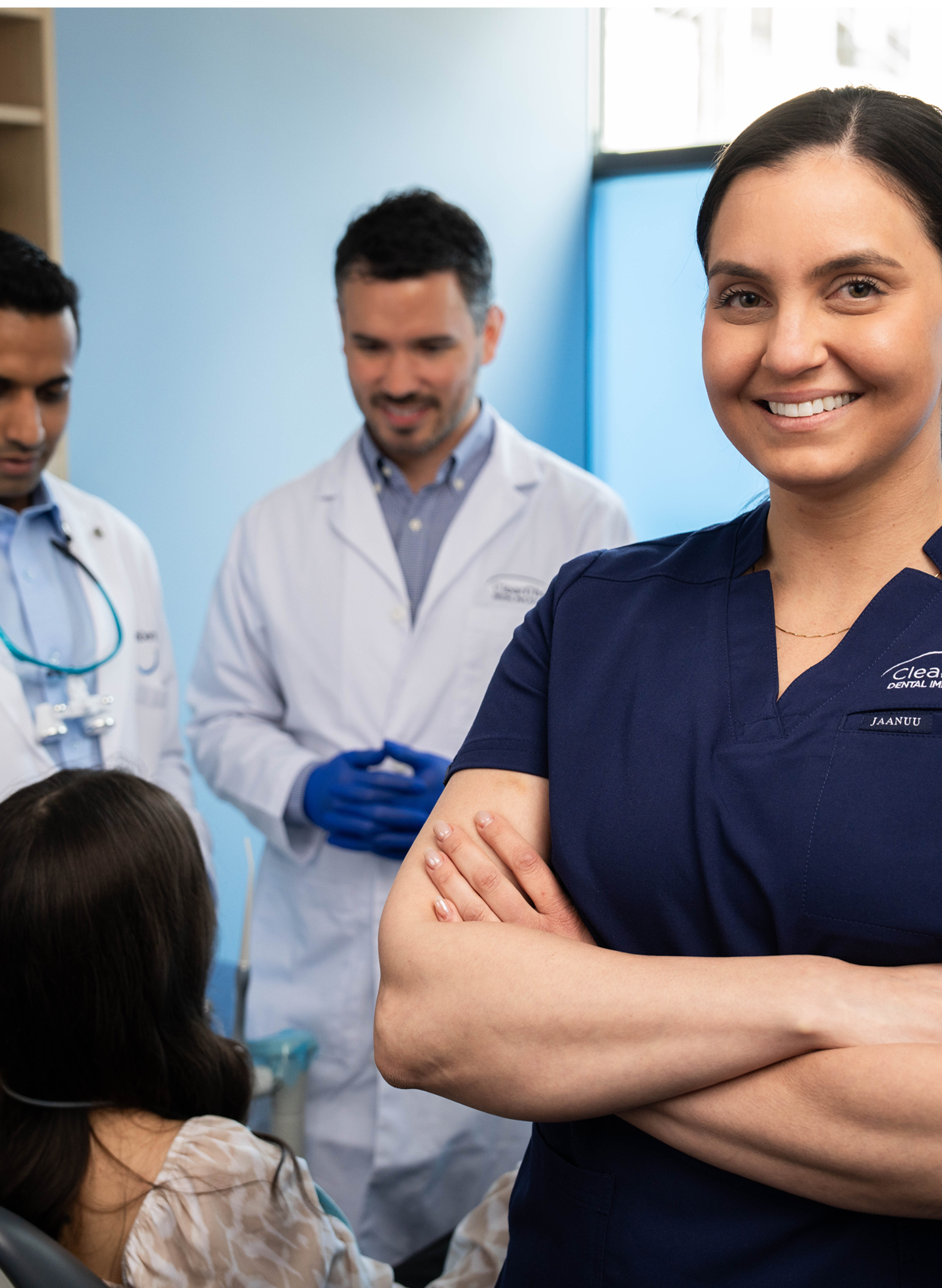 A smiling Aspen Dental professional stands confidently in the foreground while dentists consult with a patient in the background. The image represents a dental bonding before and after experience, highlighting expert care. A clean, modern office reflects a commitment to quality cosmetic dentistry.