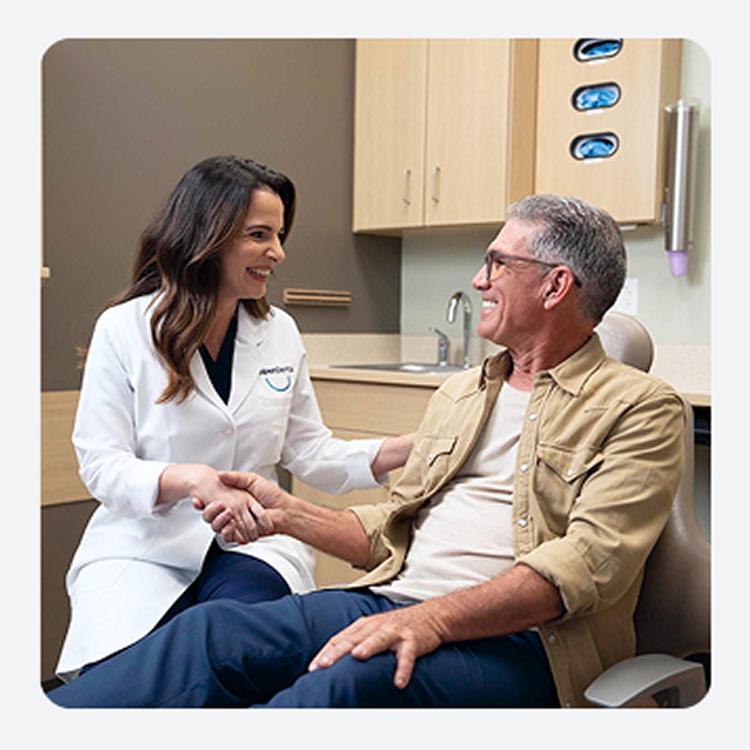 Una profesional dental de Aspen Dental sonriente con bata blanca estrecha cálidamente la mano de un paciente masculino mayor en una sala de examen dental. Parecen tener una conversación amistosa y tranquilizadora, lo que sugiere una experiencia dental positiva.