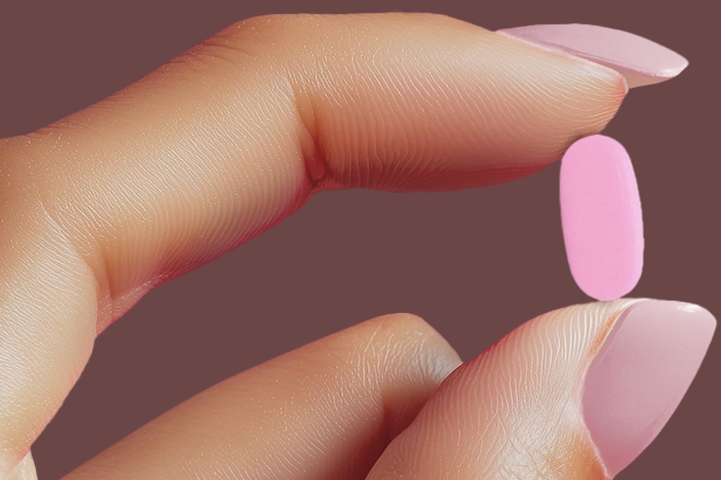 A hand with pink painted nails holds a bright pink pill against a dark background
